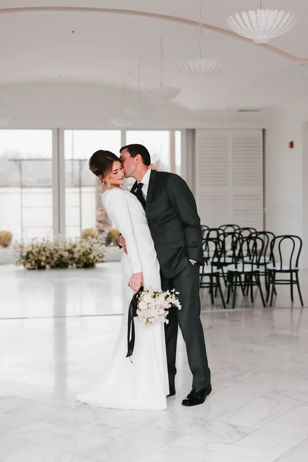 A bride and groom share a kiss in a bright, minimalist wedding venue with white walls, large windows, black chairs, and floral decorations. The bride wears a white gown holding a bouquet, and the groom wears a black suit.