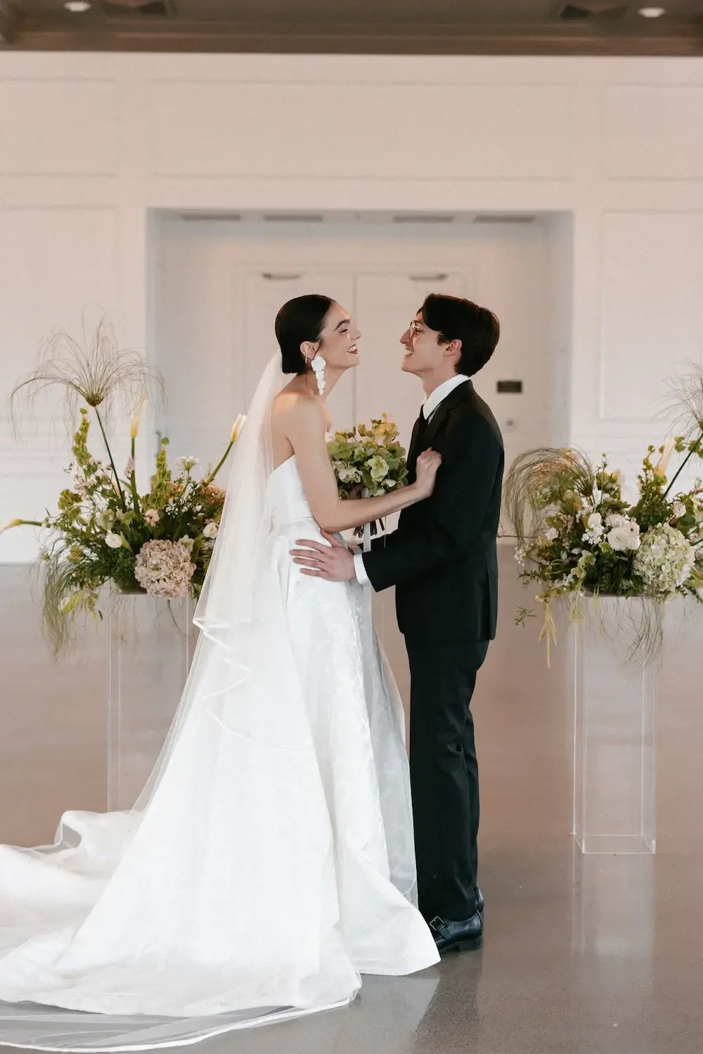 A bride and groom stand face to face, smiling at each other, during their wedding ceremony. The bride wears a white strapless wedding gown and veil, while the groom is in a black suit and tie. They are holding a bouquet, and large floral arrangements