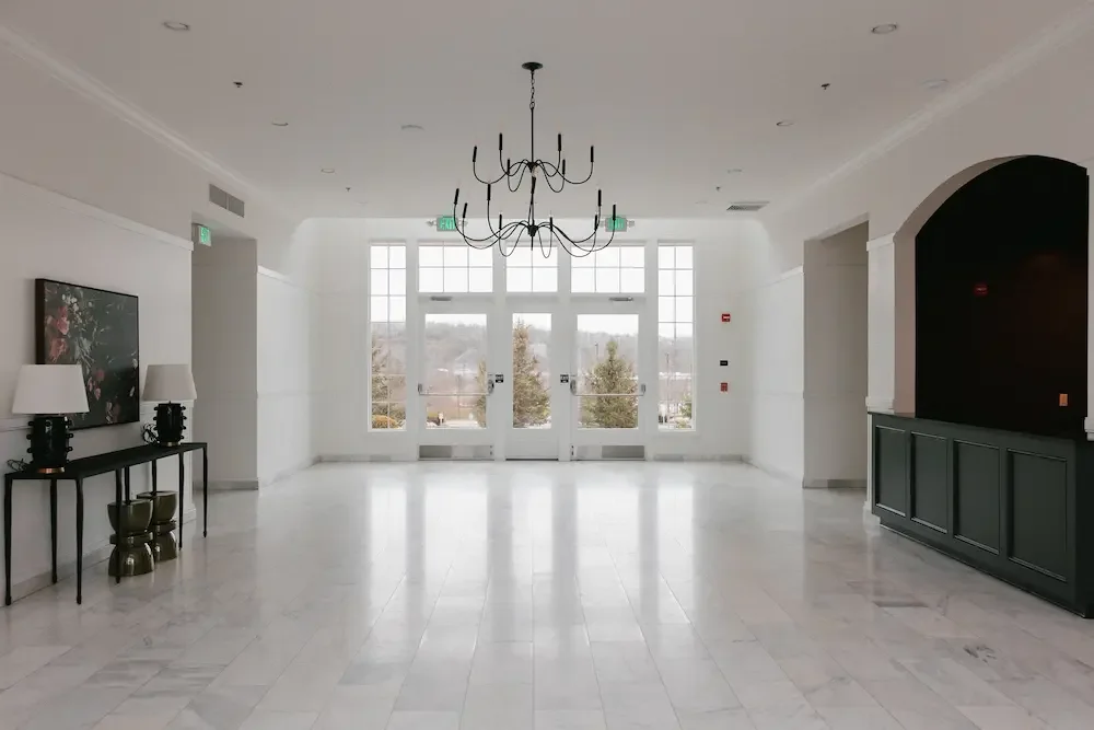 Empty, well-lit lobby with large windows, a chandelier, a console table with lamps on the left, and a dark accent wall on the right.