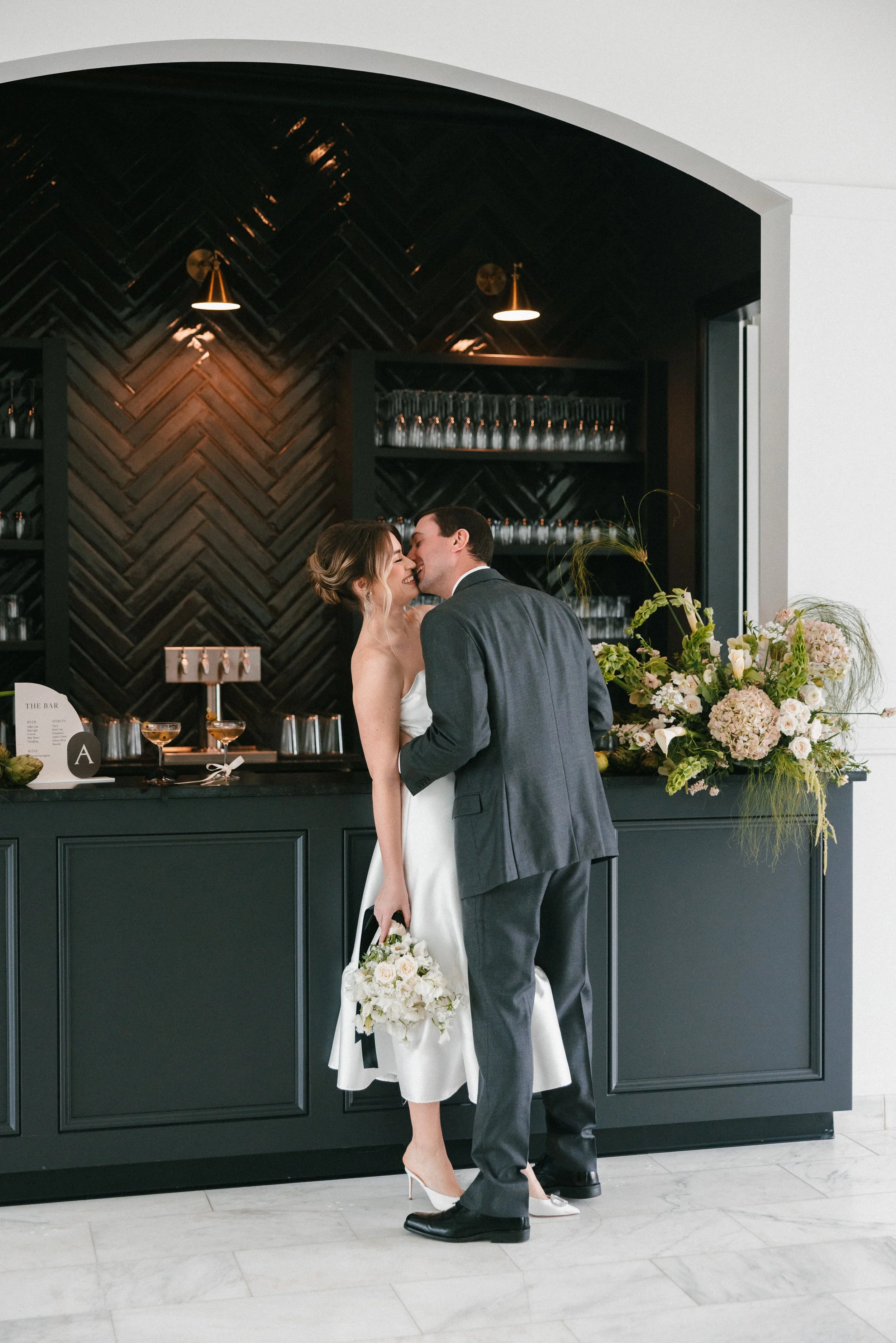 A bride and groom sharing a kiss at a wedding reception, standing in front of a dark bar with glass shelves, floral arrangements, and dim lighting.