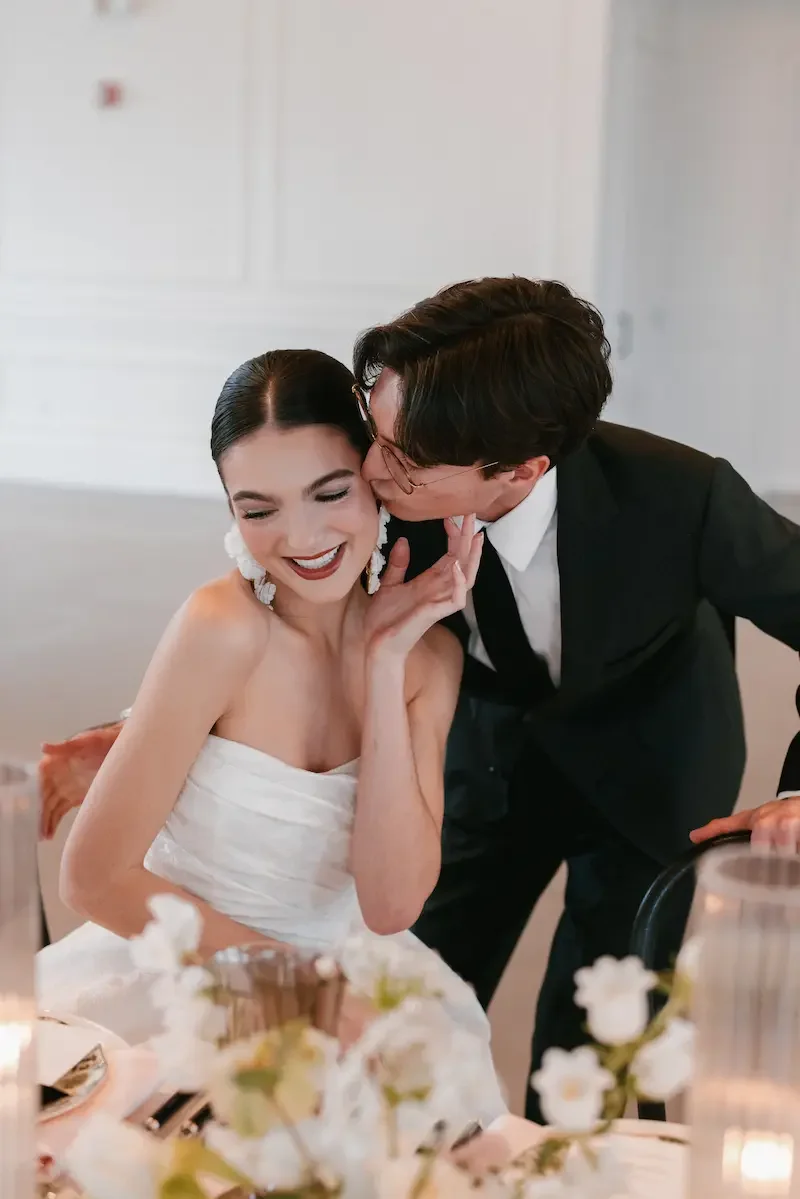 A joyful bride and groom sharing a kiss at their wedding reception, with the groom leaning in to kiss the bride's cheek, who is smiling with eyes closed, sitting at a decorated table with flowers and candles.