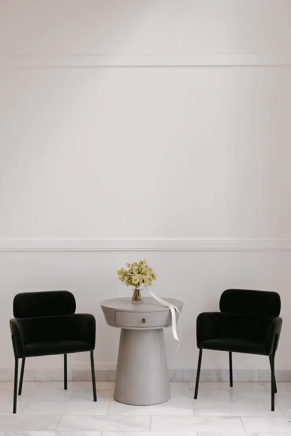 Minimalist interior with two black chairs flanking a small gray table with a drawer, holding a vase of white flowers with a white ribbon.
