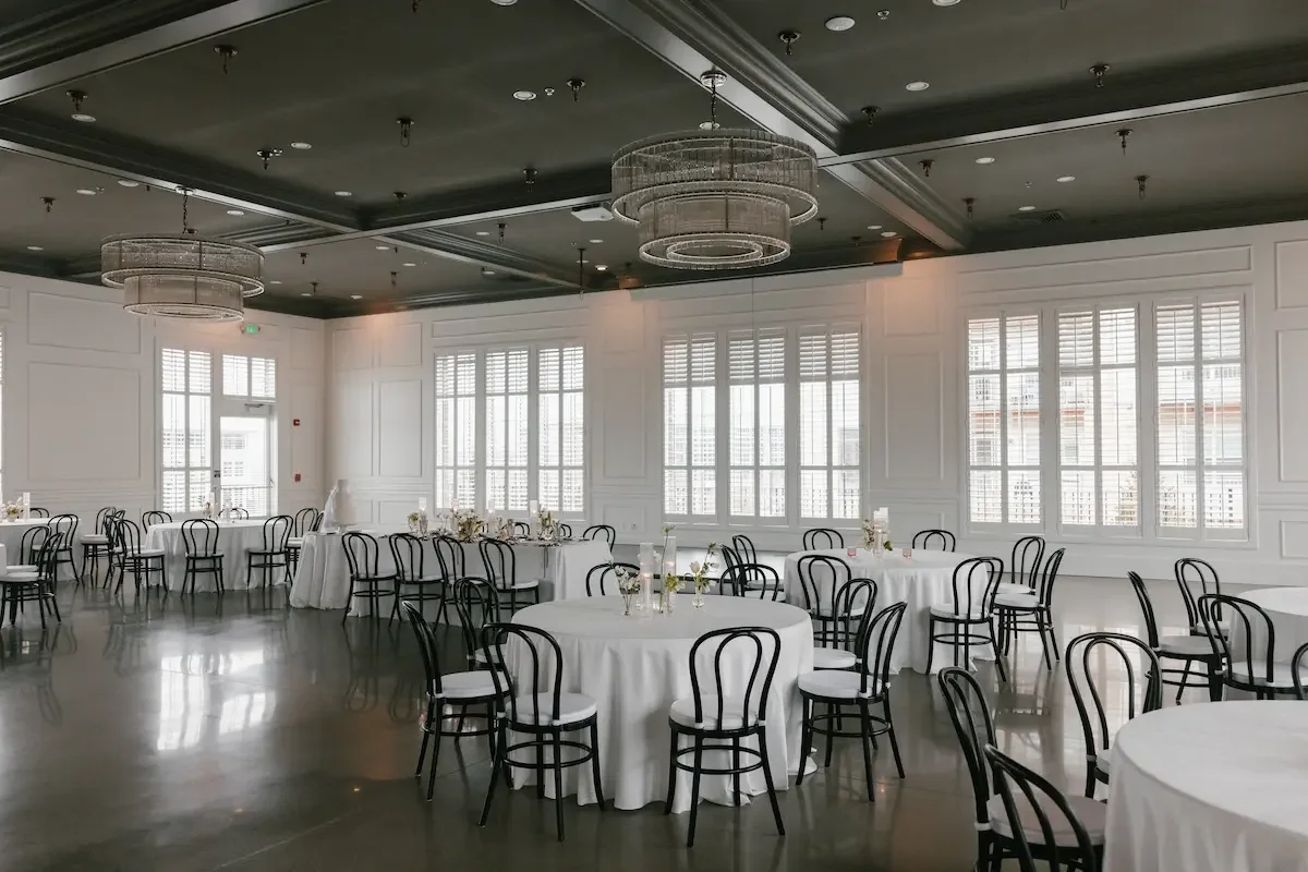 Elegant banquet hall with white walls, high windows with shutters, round tables with white tablecloths, black chairs, and modern chandeliers hanging from a dark ceiling.
