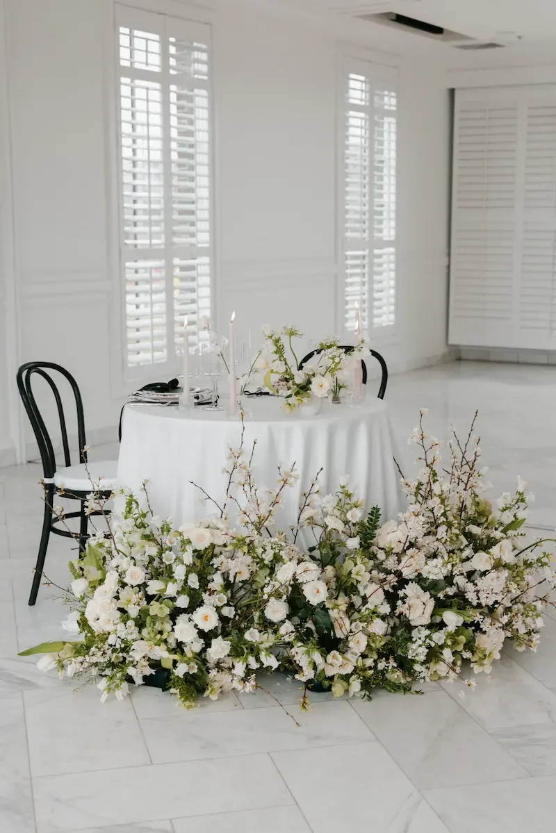 Elegant round table decorated with white flowers and candles, set in a bright white room with white shutters and marble floor.
