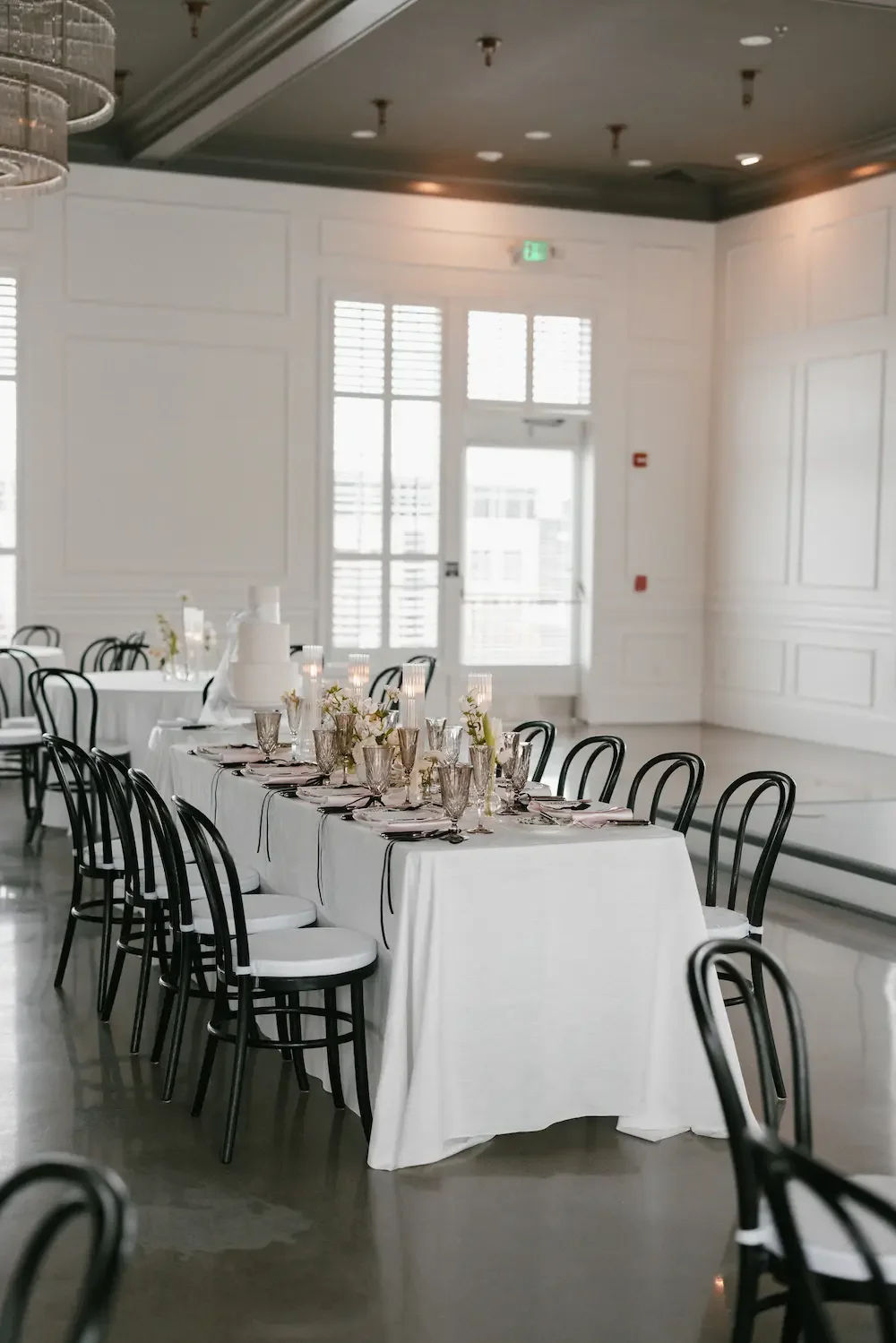 Elegant banquet table setup in a bright, modern room with white walls and large windows, black chairs, white tablecloth, floral centerpieces, and glassware.