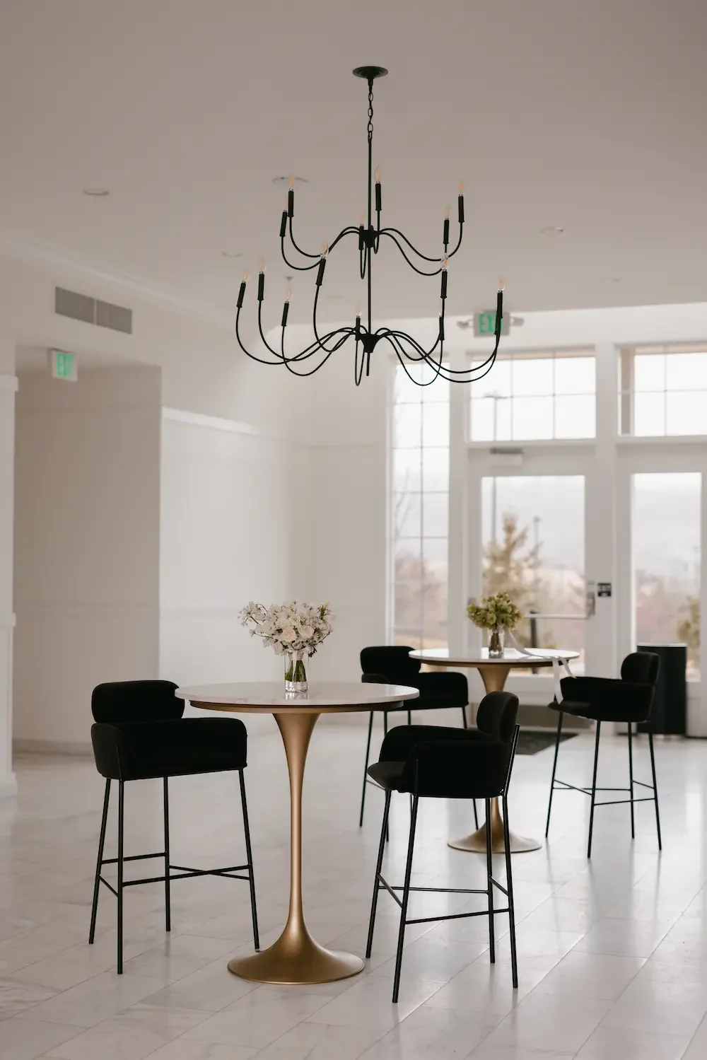 Modern interior space with black bar-height chairs around two round tables with white tabletops and floral centerpieces, large windows, and a black chandelier hanging from the ceiling.