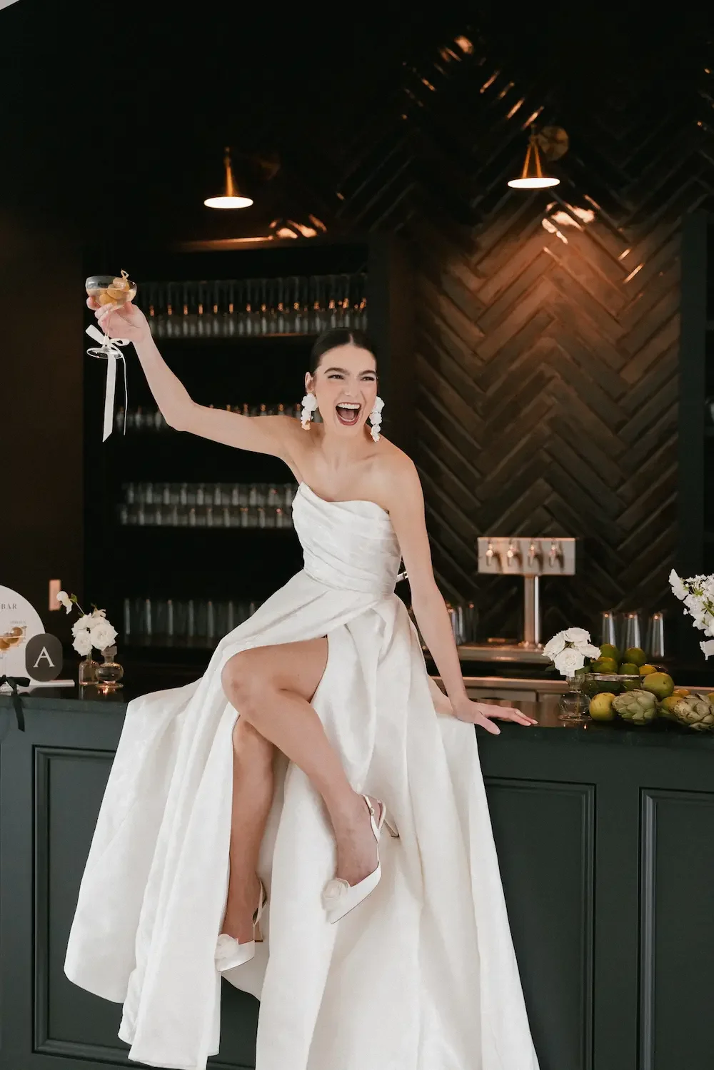 Joyful bride in a strapless white wedding gown, sitting on a bar counter, raising a cocktail glass, and smiling broadly, in a modern bar setting with dark wood accents.