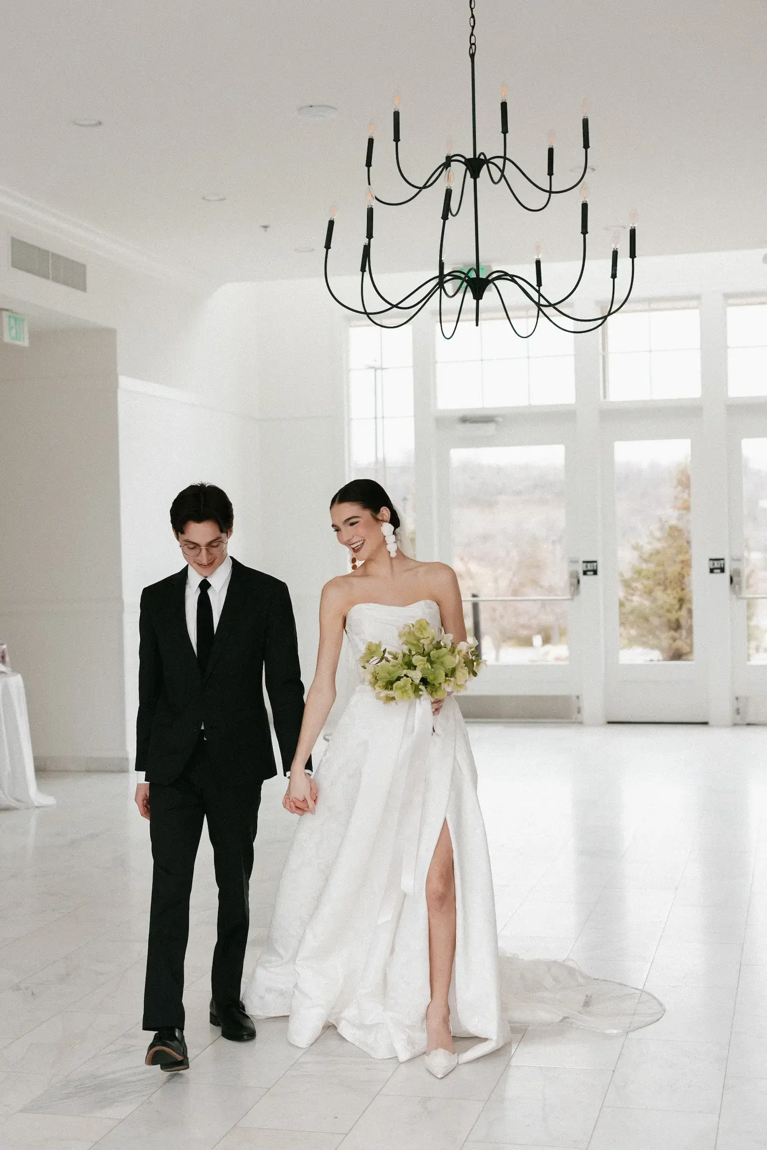 A bride and groom walking hand in hand inside a bright, modern venue with large windows. The bride is in a white strapless wedding gown holding a bouquet, and the groom is in a black suit with a white shirt and black tie. An ornate chandelier hangs from the ceiling.