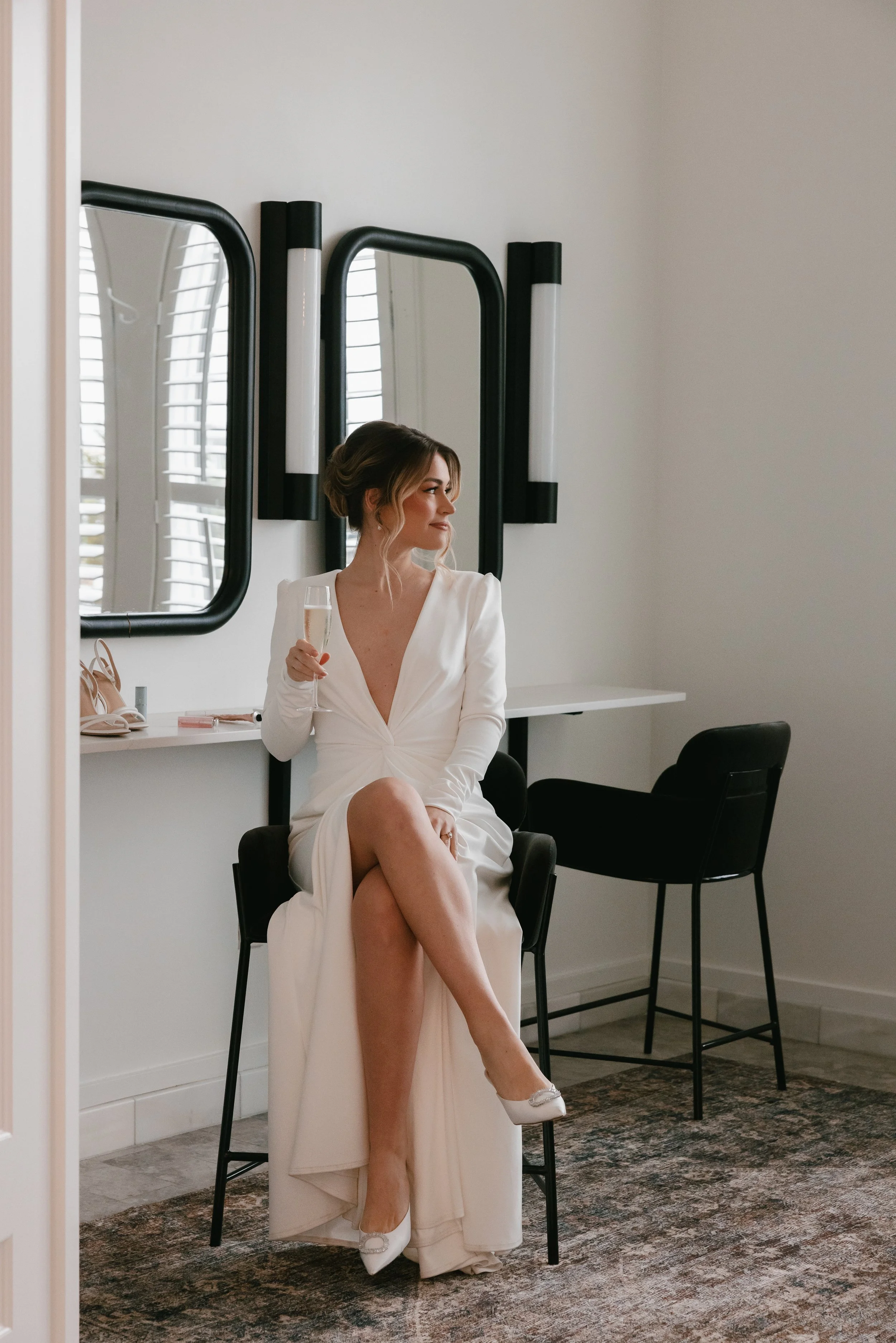 A woman in a white dress sitting on a black chair while holding a glass of champagne in a room with white walls, black-framed mirrors, and a patterned rug.