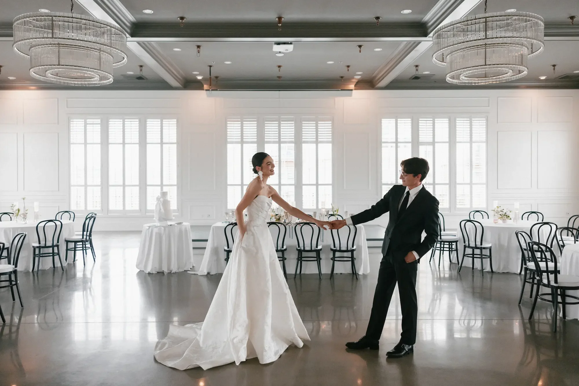 A bride and groom dancing in a decorated banquet hall with tables and chairs, chandelier lighting, and large windows.