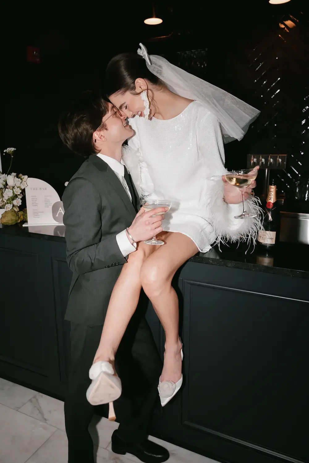 A bride and groom celebrating at a bar, with the bride sitting on the bar counter holding a champagne glass, and the groom holding her leg. Both are smiling and dressed in wedding attire, with champagne glasses in hand.