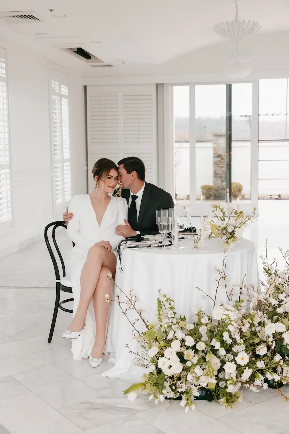 A bride and groom sitting closely together at a wedding reception, with the groom whispering into the bride's ear. The table has a white tablecloth, glasses, and floral arrangements, and the room is filled with natural light and bright decor.