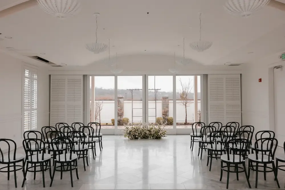 Empty wedding ceremony room with black chairs arranged in rows, large windows, white floral arrangement at the front, and outdoor landscape view.