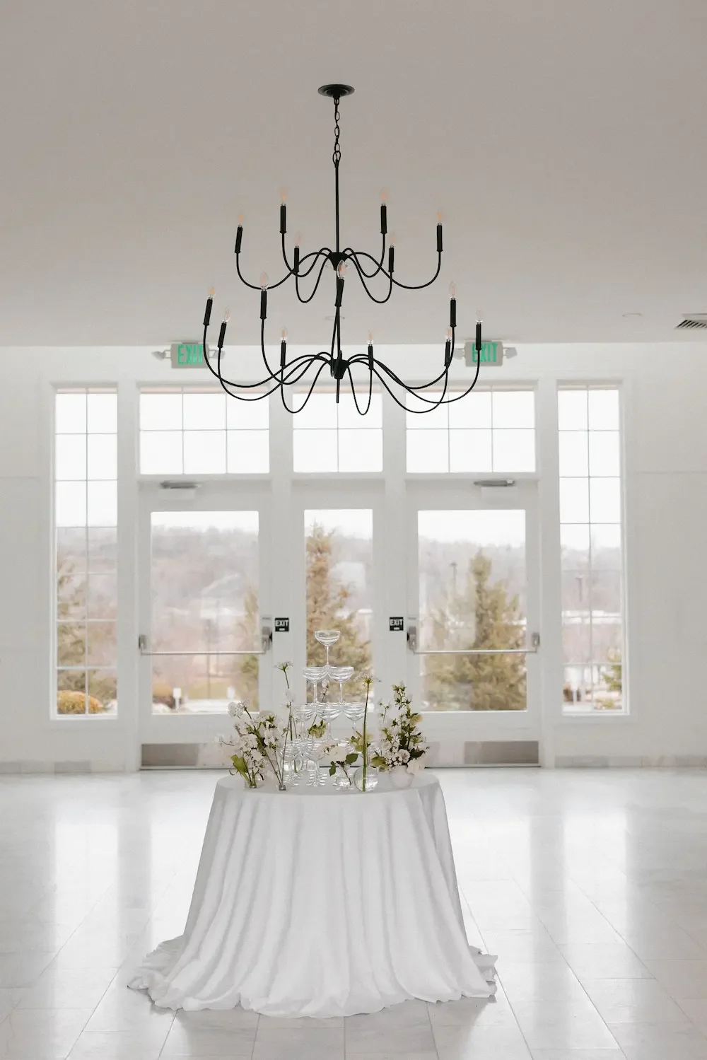 Elegant white room with large windows, a black chandelier, and a round table with a white tablecloth, floral arrangements, and glassware.