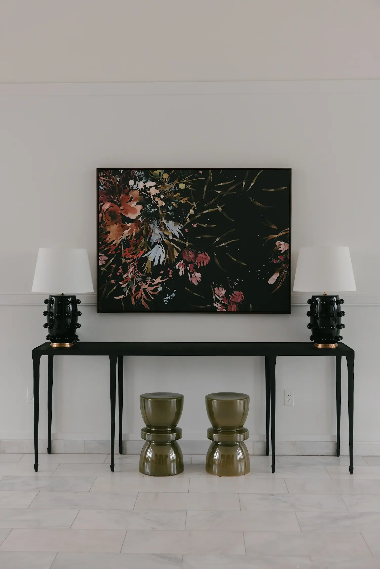 A modern living room with a white wall, two table lamps on black side tables, a black table with gold accents, two gold stools, a painting of flowers on the wall, and white marble flooring.