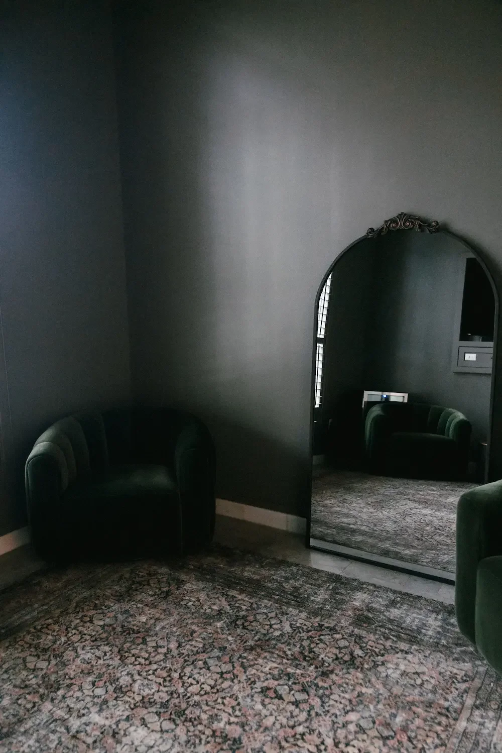 A dark room with black walls and a black carpet. There is a large, ornate mirror reflecting a green velvet armchair and a window with blinds.