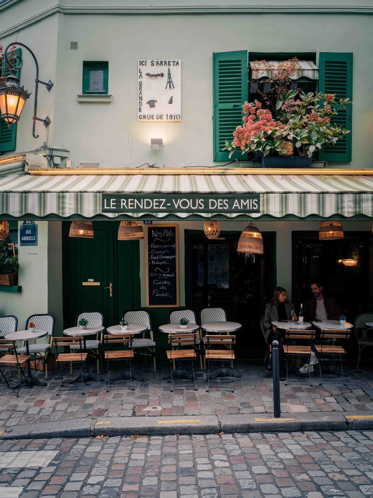 French café with outdoor seating, striped awning, and green shutters. Two people sit at a table, and a chalkboard menu displays food options. Decorative lanterns hang from above.