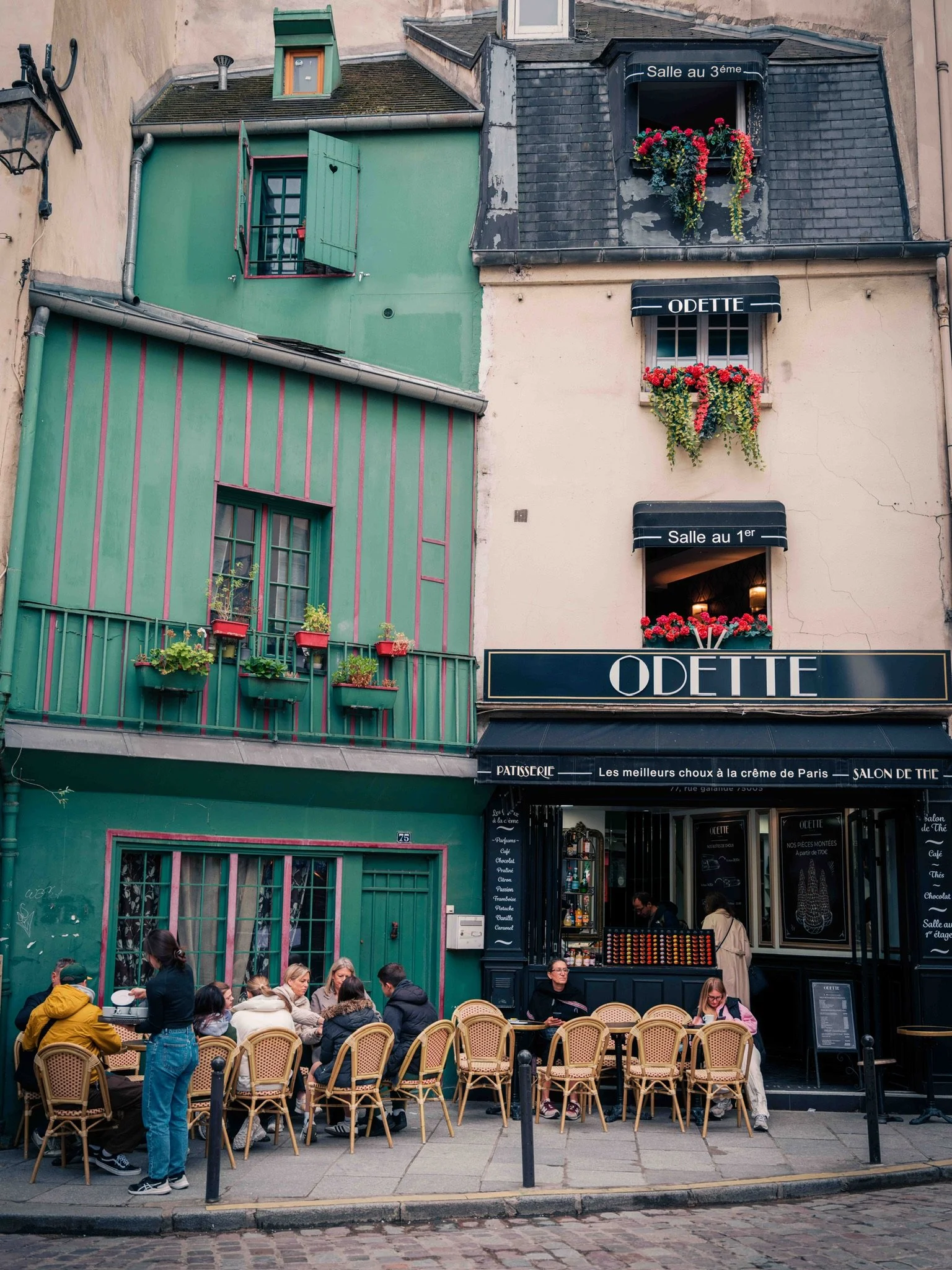 Outdoor scene of a cafe in Paris, France, with people sitting at tables on the sidewalk. The building has multiple floors with colorful window shutters, flower boxes, and signs indicating rooms and a pastry shop named Odette.