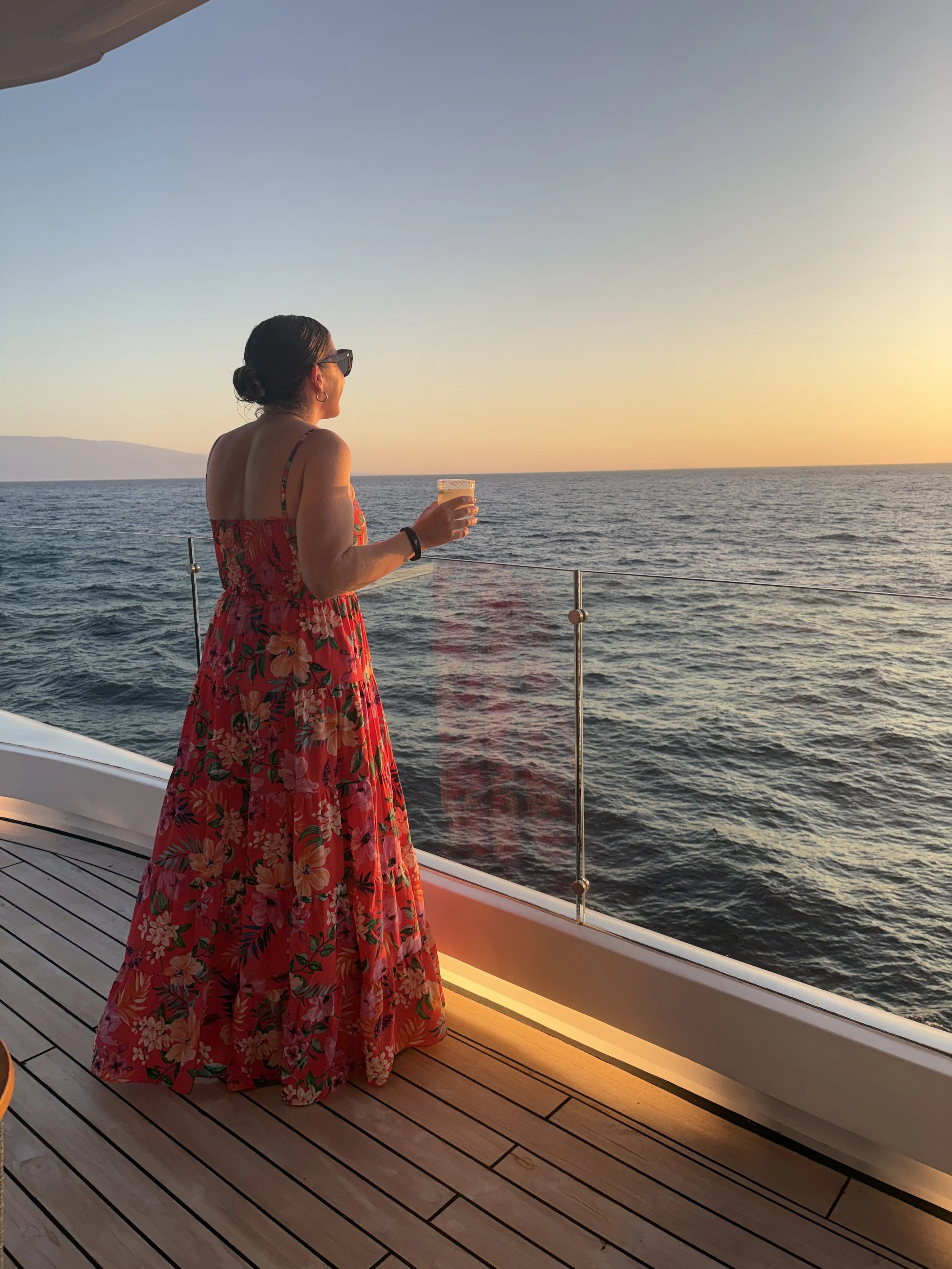 A woman in a floral red dress and sunglasses holding a drink on a boat during sunset, looking out at the ocean.