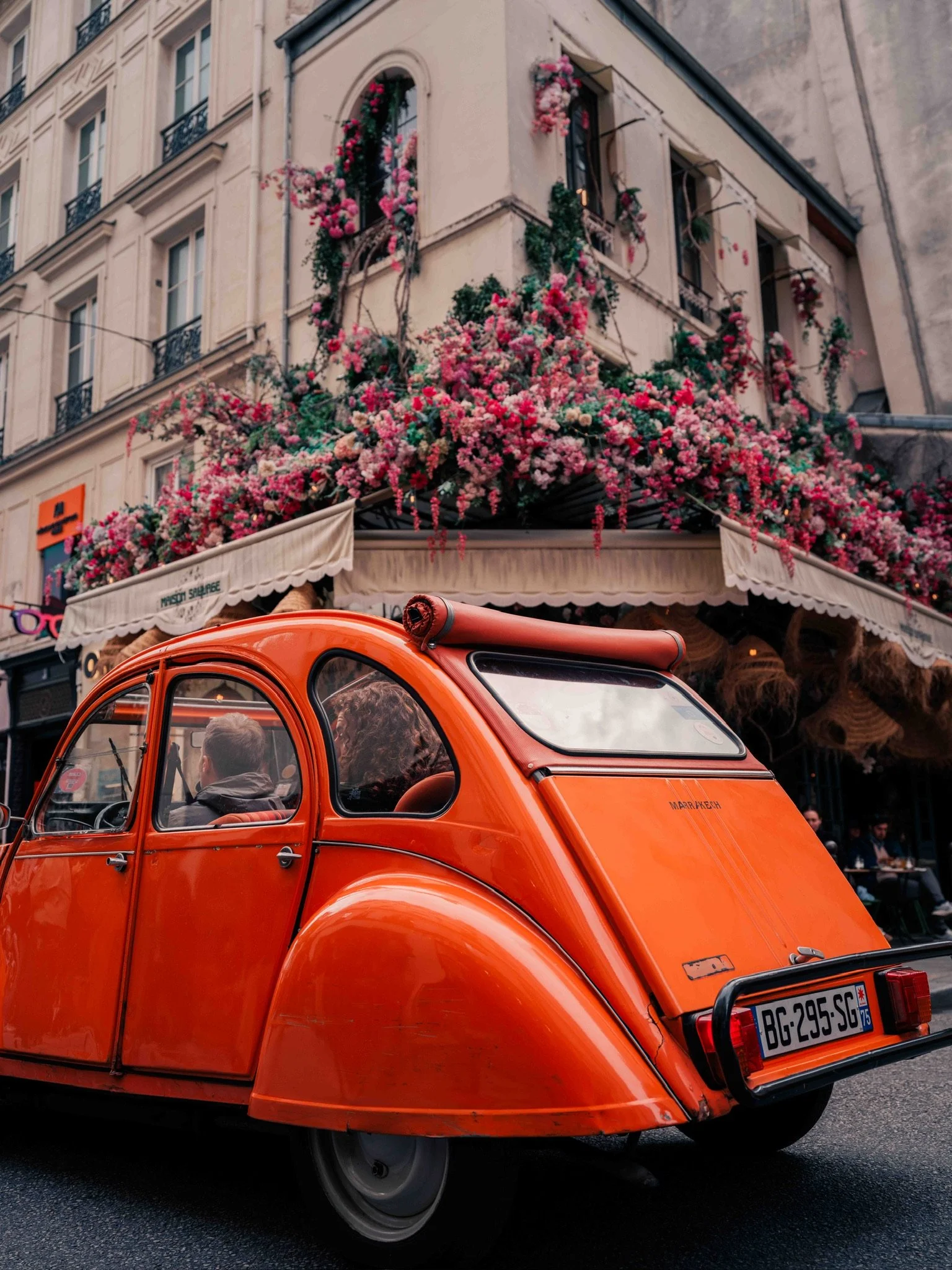 An orange vintage car parked in front of a building decorated with pink and red flowers.