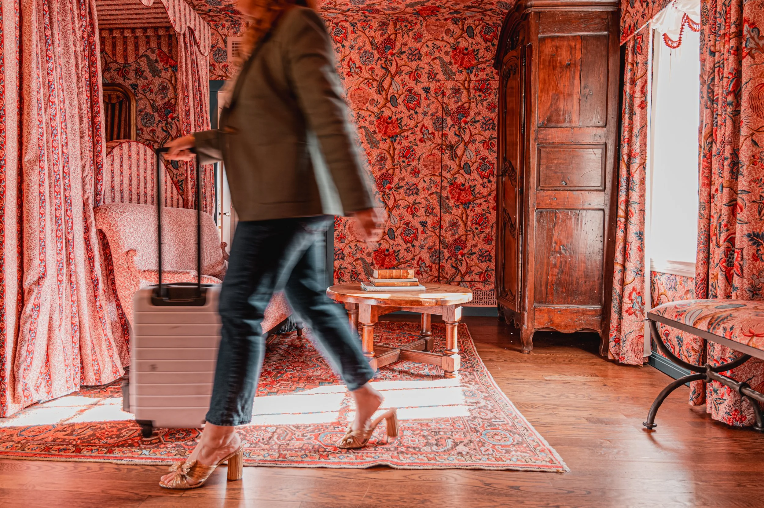 Woman walking through a room with red patterned wallpaper, carrying a rolling suitcase, wearing high heels and dark pants. The room has vintage furniture, including a wooden wardrobe, a round table with books, and floral curtains.