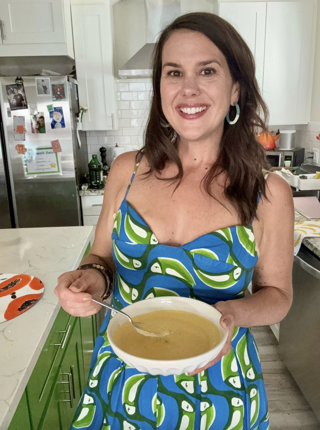 A brown haired woman in a green and blue patterned dress, stands holding a bowl of yellow soup in front of her