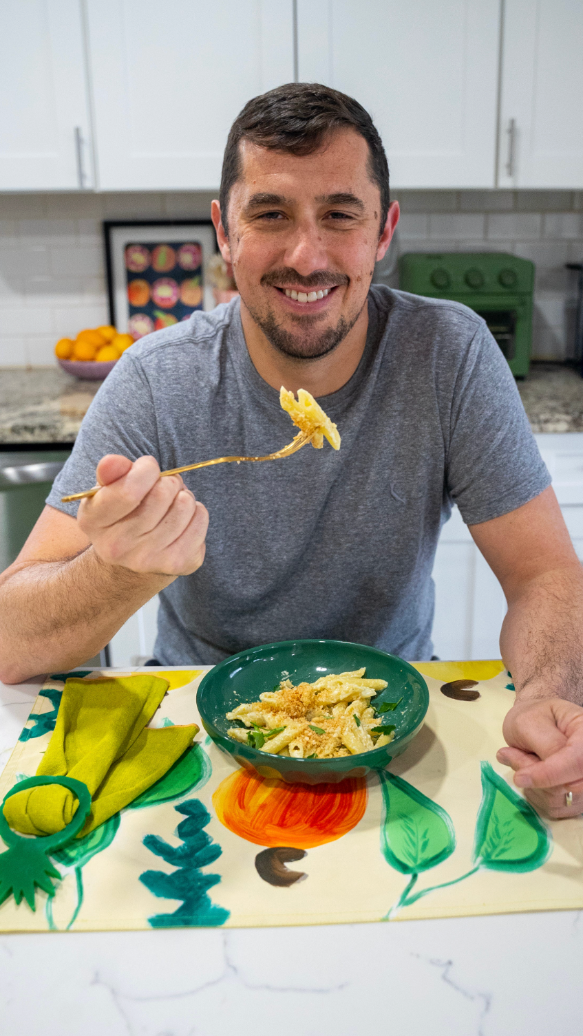 Smiling brown haired man sits at a marble countertop with a green bowl of mac and cheese in front of him, holding up a bite on his fork