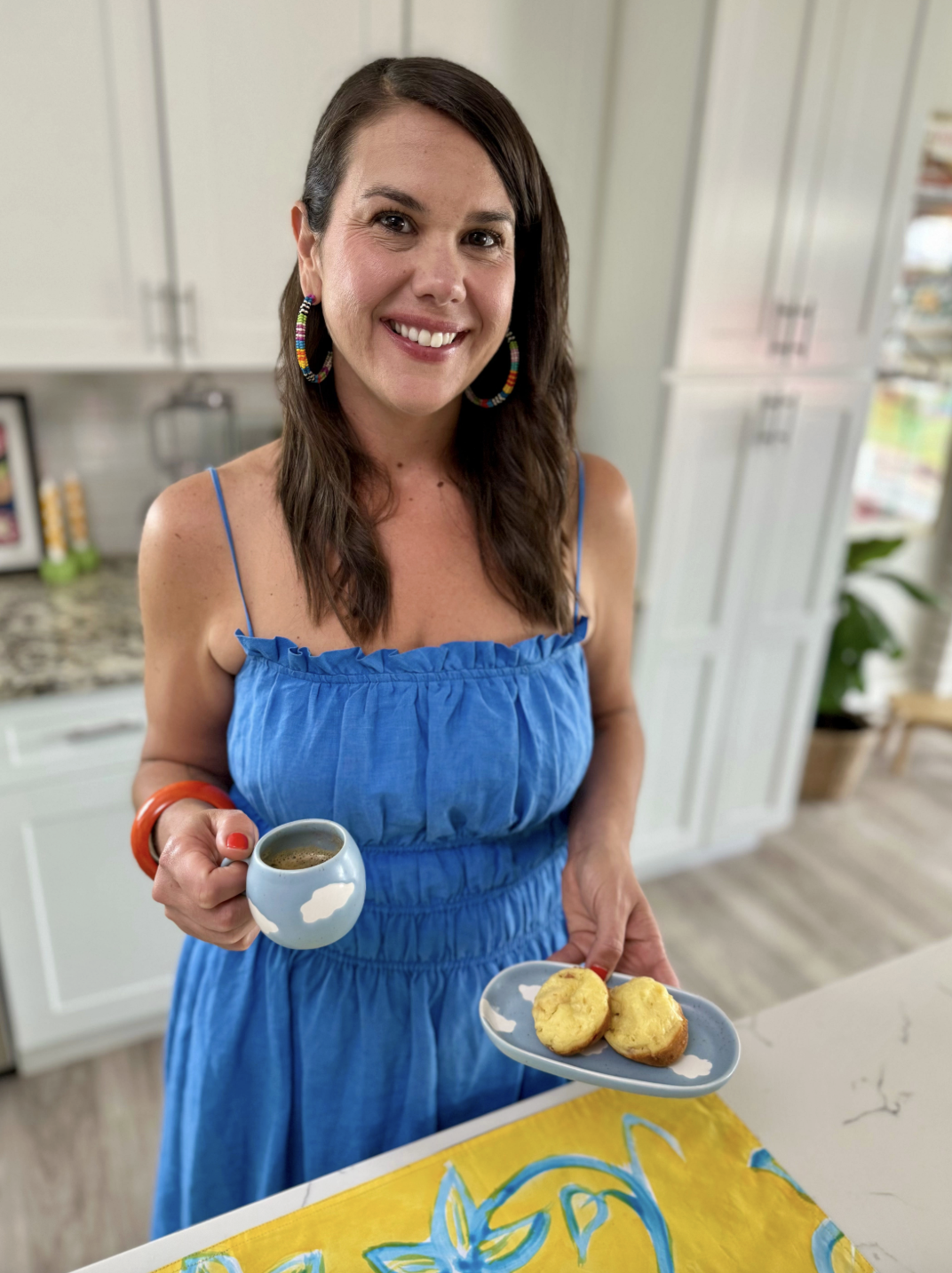 A brown-haired woman in a blue sundress smiles while holding a small blue espresso cup in one hand and a plate of two tapioca breakfast cups in the other.