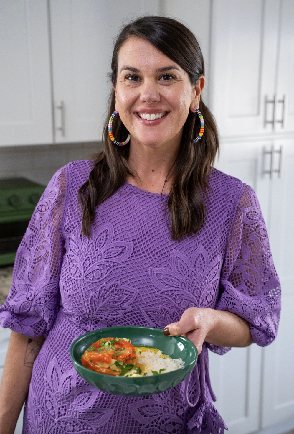 A smiling brown-haired woman in a pruple dress, holding out a green bowl filled with rice and a halibut, green papaya moqueca and tomato mixture topped with cilantro.