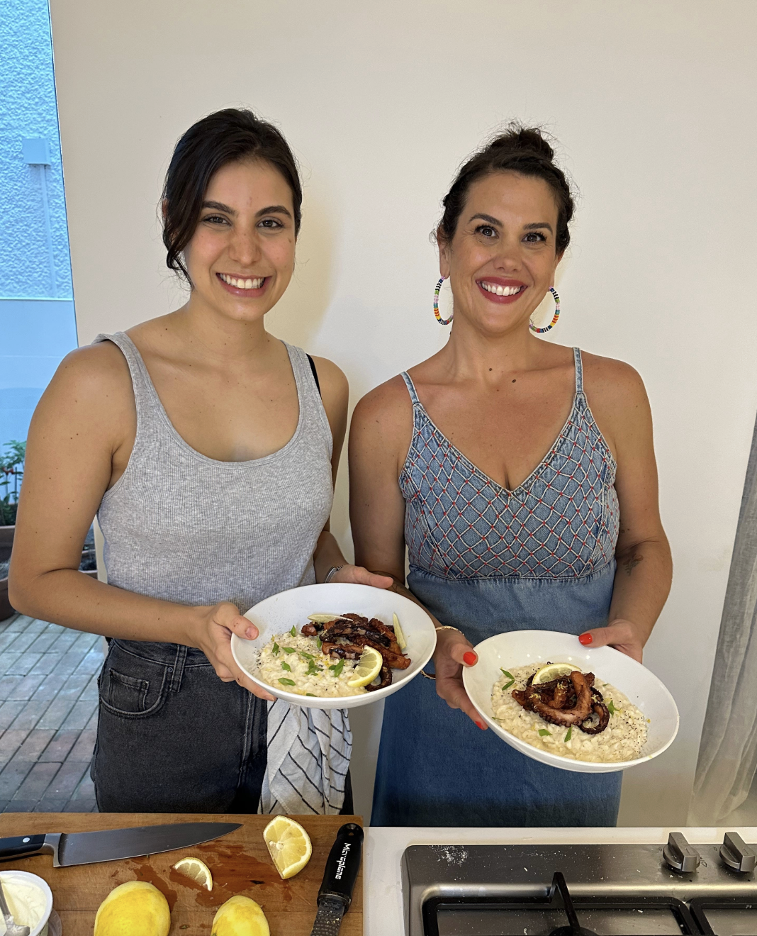 The image shows two brown-haired, brown-eyed women standing next to each other, smiling at the camera while holding white bowls that contain grilled octopus on top of a lemon risotto.