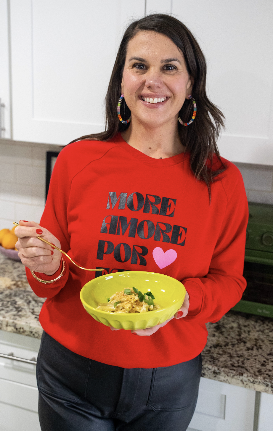 Smiling, brown-haired woman in a red sweater holds a bowl of mac and cheese in one hand with a gold form in the other