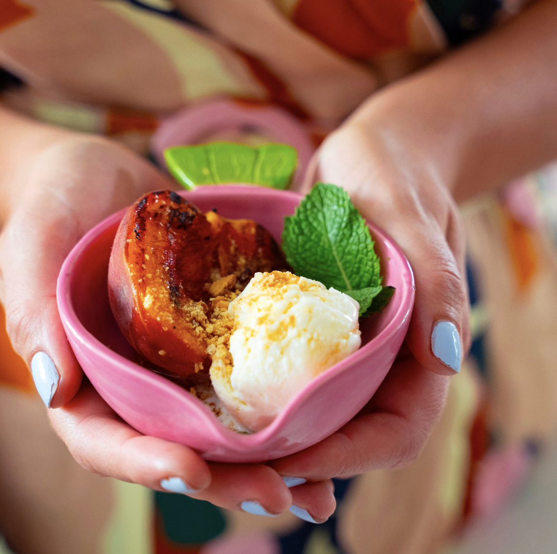 A scoop of vanilla ice cream, half a grilled peach topped with graham cracker crumbs and mint leaves.