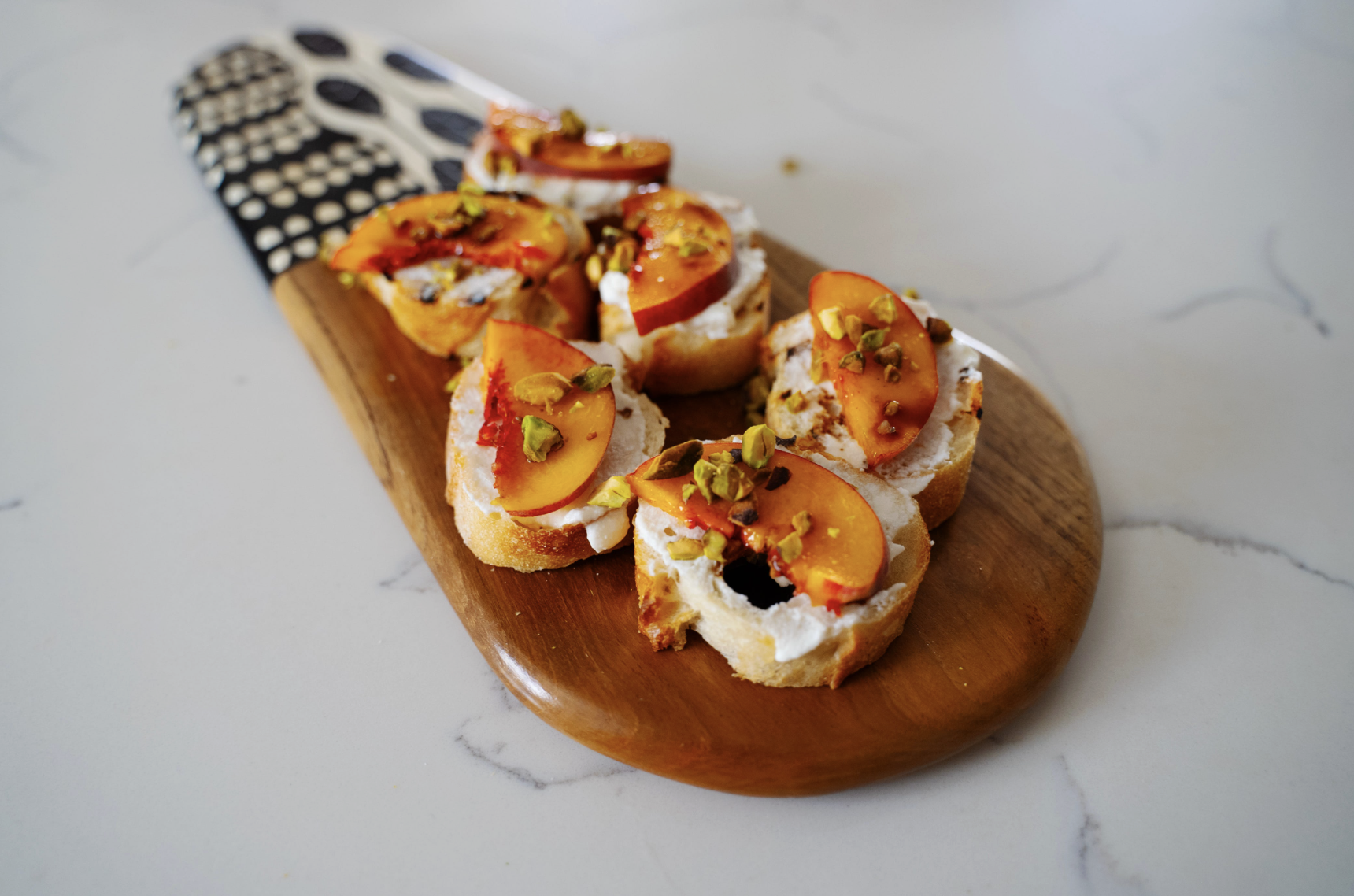 A wodden serving platter of ricotta, sliced peach, and pistachio crostinis