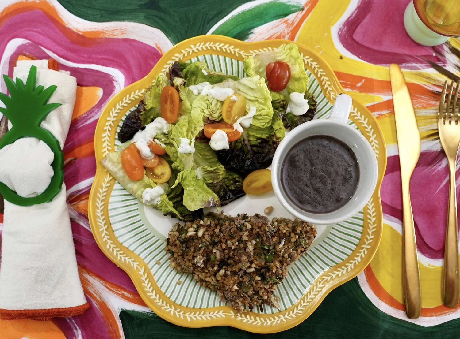 The images shows a plate serving oof kibe, a white cip of black beans and a green salad with cherry tomatoes.