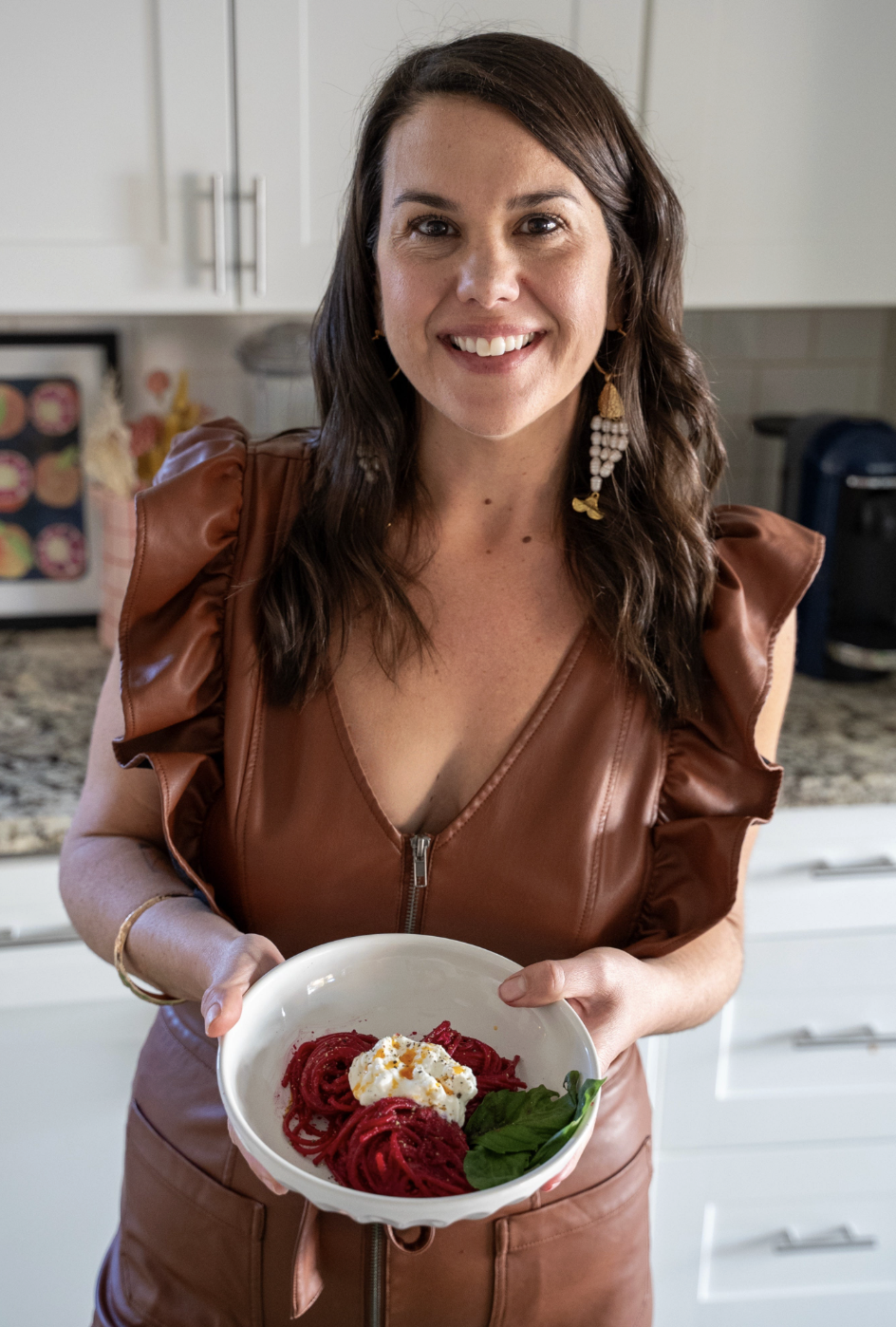 A brown hiared woman in a brown leather dress, smiles an dholds out a white bowl of red beet pasta, topped with burrata, chili oil and basil leaves.