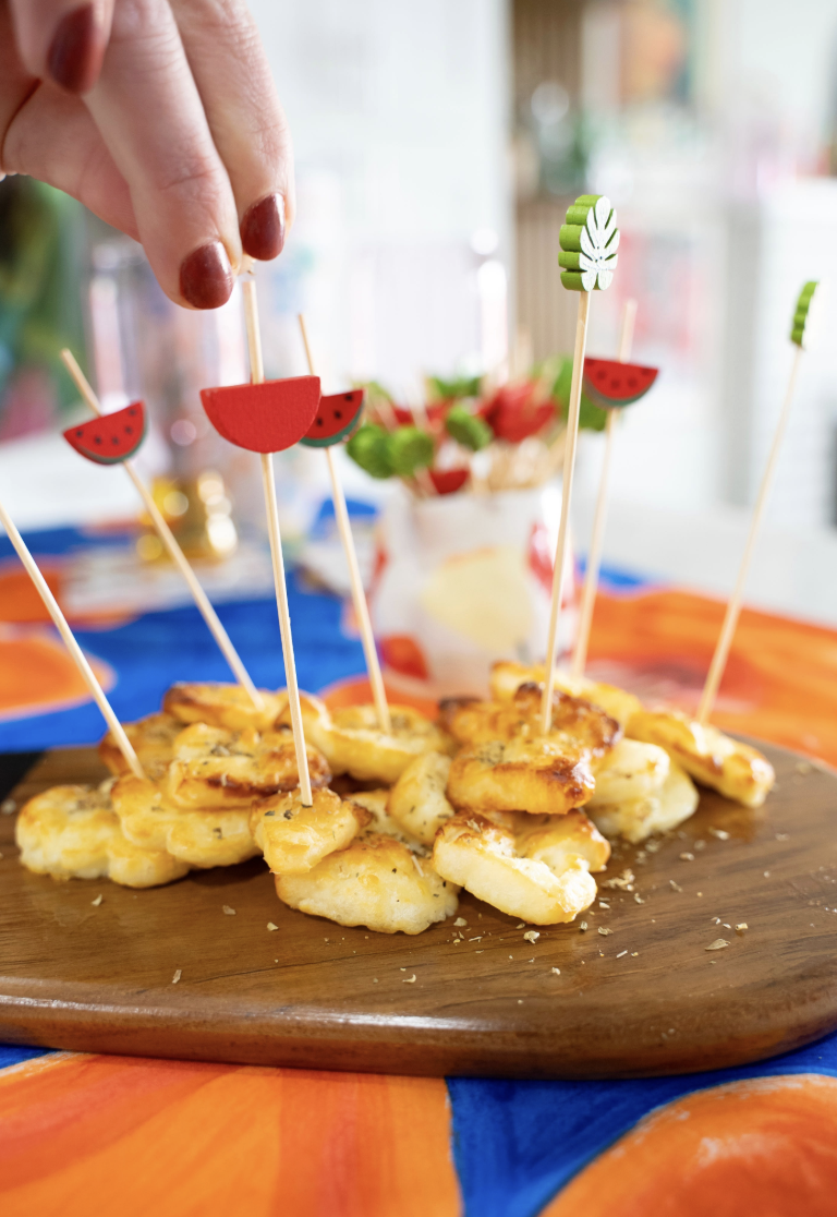 The image depicts a hand picking up a skewer with decorative watermelon slice from a wooden board with stacked cheese appetizers, with more skewers in the background.