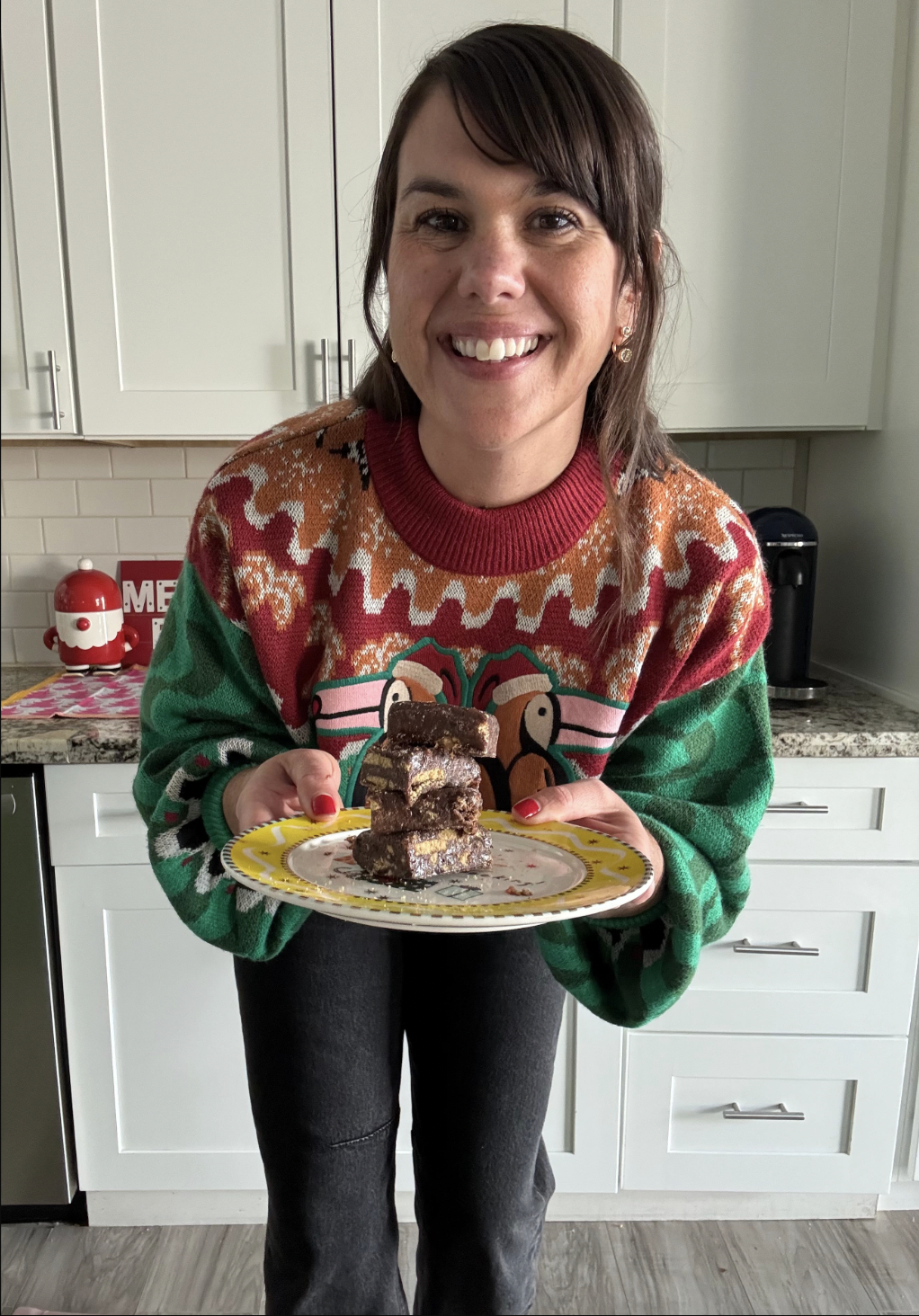 A smiling woman in a holiday sweater showing a plate of fudgy palha italiana squares to the camera.
