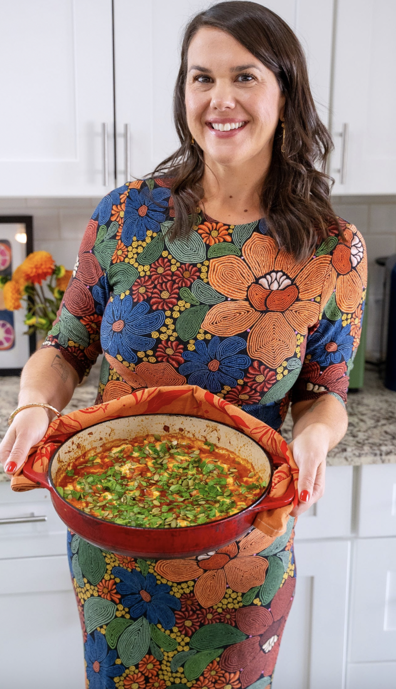 The image depicts a brown haired woman, smiling and holidng a large red skillet filled with red sauce and topped with green herbs and pepitas