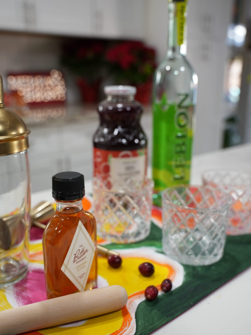 The image shows a small amber bottle of cardamom bitters infront of two cocktail glasses, a bottle of cranberry juice and a bottle of cachaca