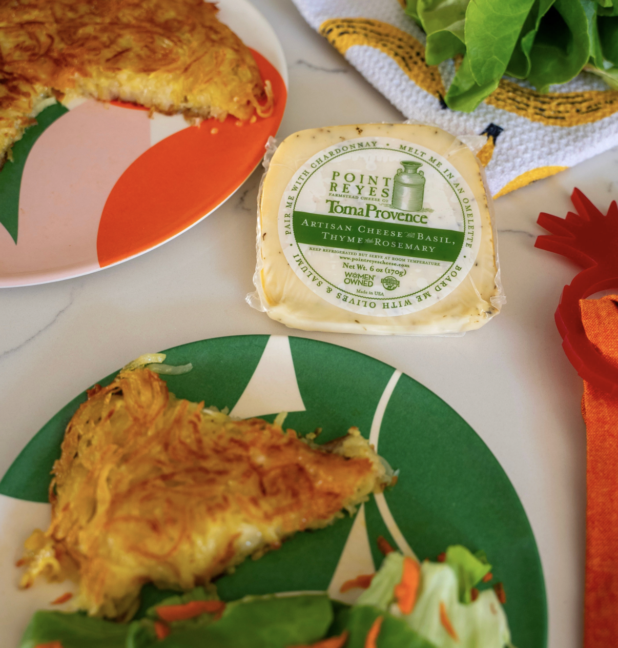 A top view of a countertop showing a serving platter of potato rosti next to a block of Point Reyes Toma Provence's Basil, Thyme and rosemary Cheese, with a plate of green salad served alongside a slice of potato rosti