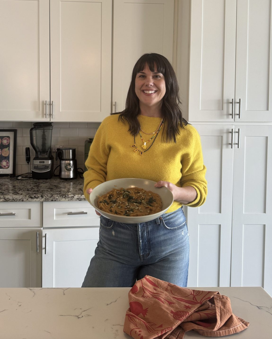 A brown haired woman in a yellow sweater and jeans, standing in front of white cabinets holding a large bowl of roasted butternut squash mash, topped with pumpkins seeds and sage
