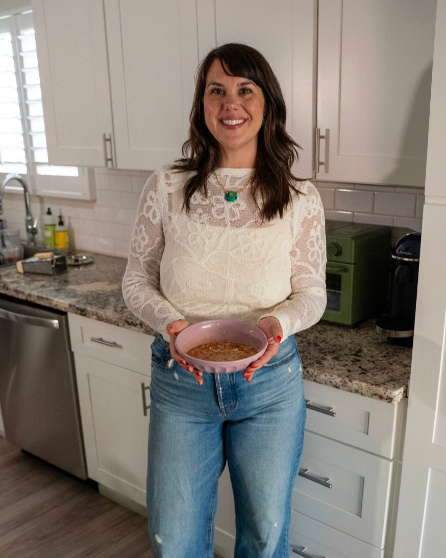 A brown haired woman in a white top and jeans holding out a bowl of Pasta e Fagioli