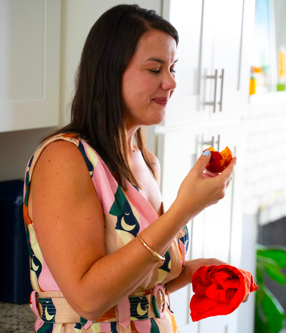The side view f a woman in a multicolored dress smiling at a bite she has just taken out of a peach