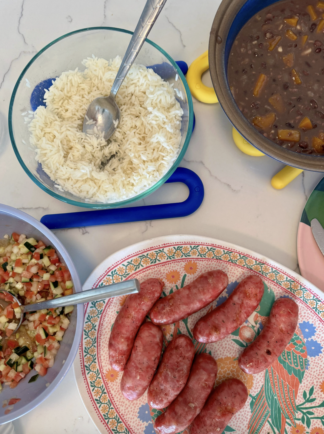 An overhead view of a table setting that has a platter of linguica, a bowl of rice, a bowl of salsa and a pot containing the vegan fall feijoada
