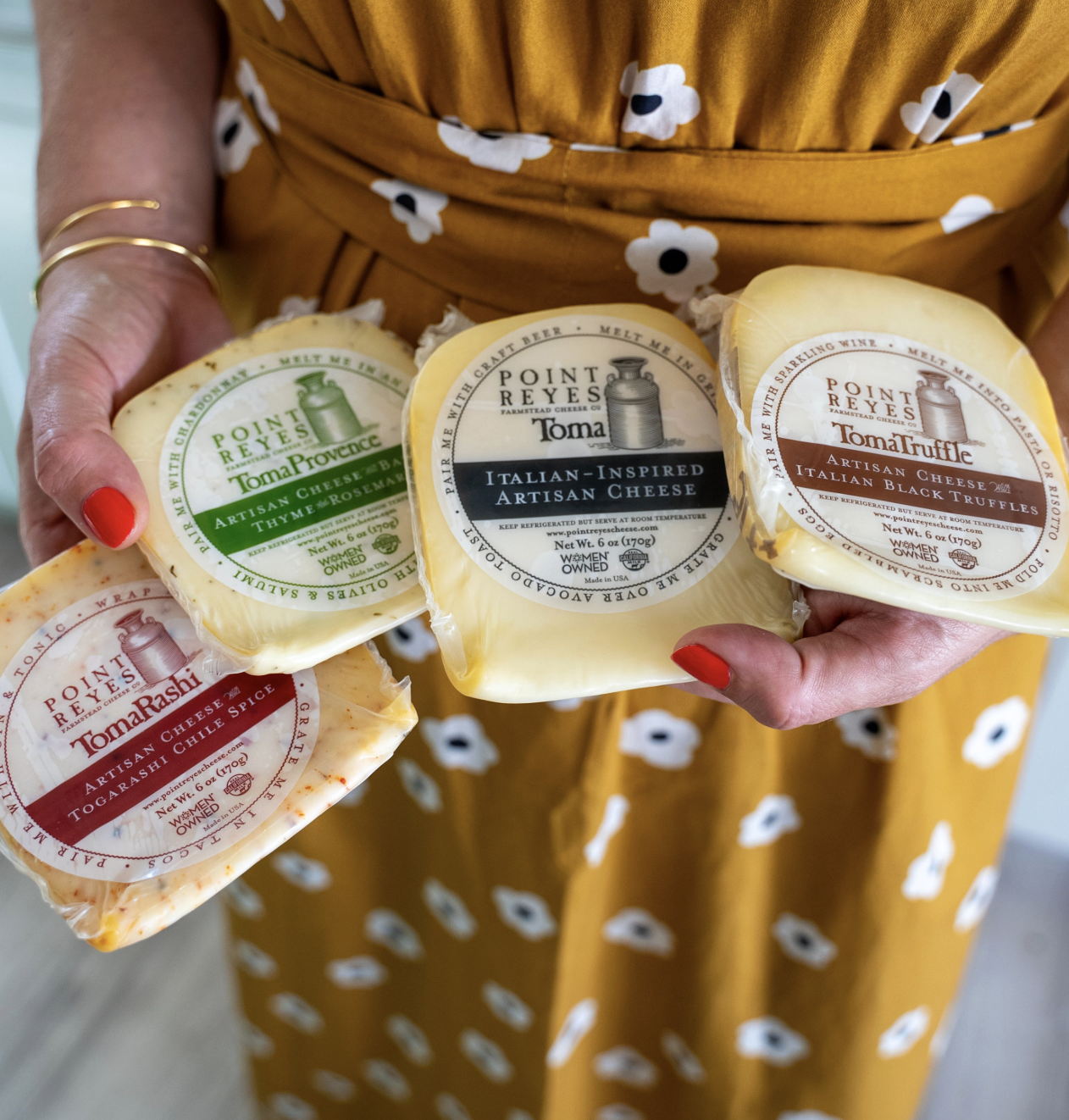 Four Point Reyes cheese being held up. From left to right, there is Toma Rashi, Toma Provence, Toma, and Toma Truffle varieties.