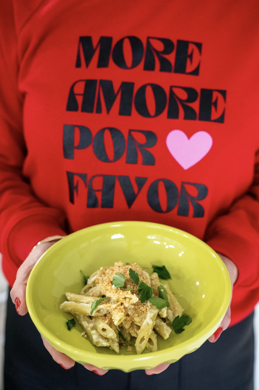 A person in a red sweater holding out a  bowl of mac and cheese penne pasta, topped with breadcrumbs and parsley