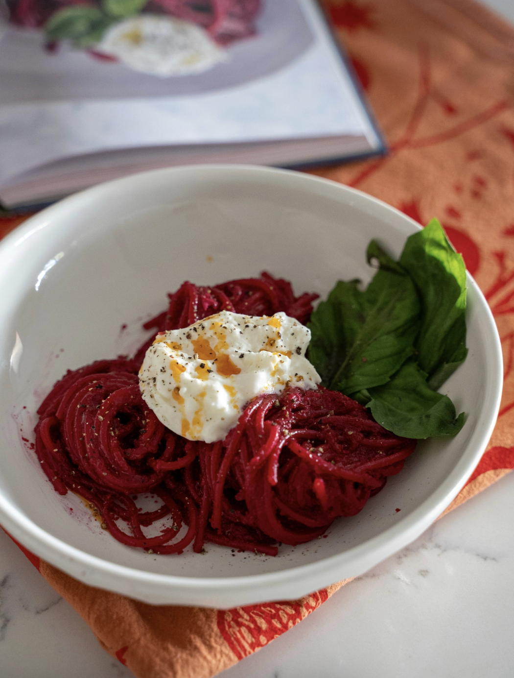 A white bowl of bright red pasta topped with burrata, chili oil and basil leaves