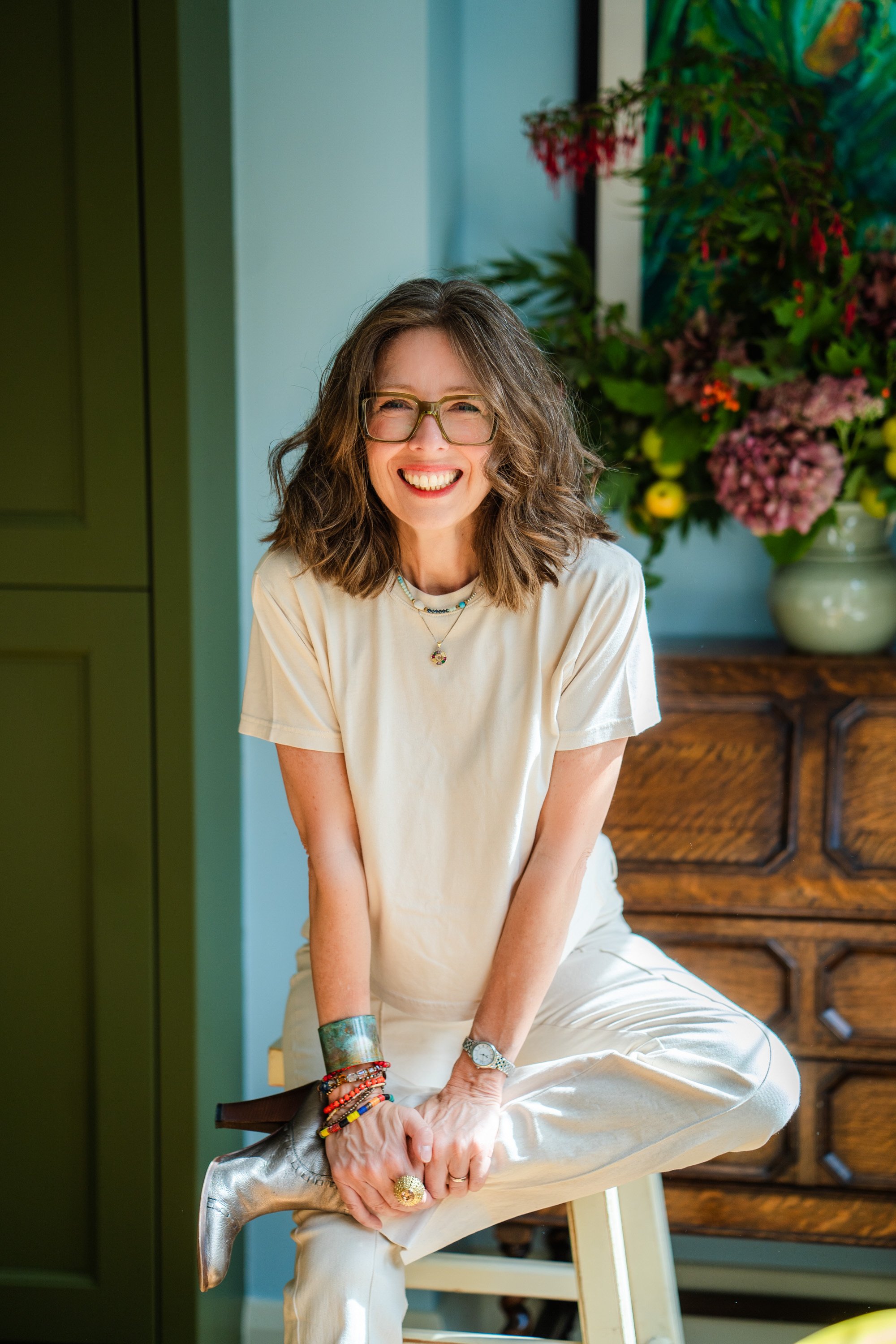 A woman with curly brown hair and green glasses laughing, sitting on a stool. She is wearing a cream t-shirt, cream jeans and silver boots. Behind her is a pale blue wall, green cupboard, antique furniture, and colourful flowers.