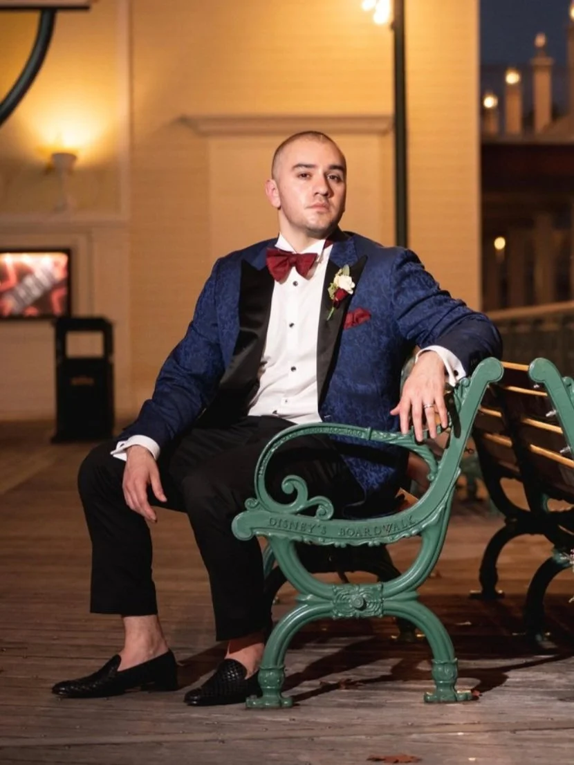 A man in a tuxedo with a blue jacket, red bow tie, and boutonniere, sitting on a park bench at night outside a building with warm lighting.