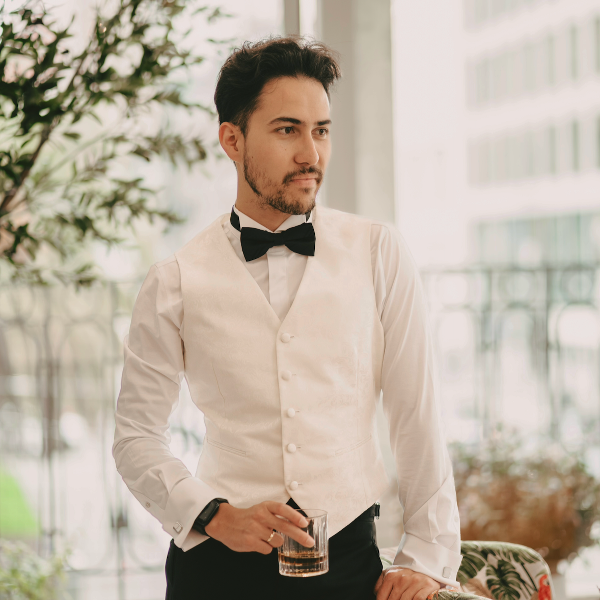 Man in a tuxedo with a black bowtie and a cream vest holding a glass of whiskey, standing in a bright indoor space with plants and large windows.